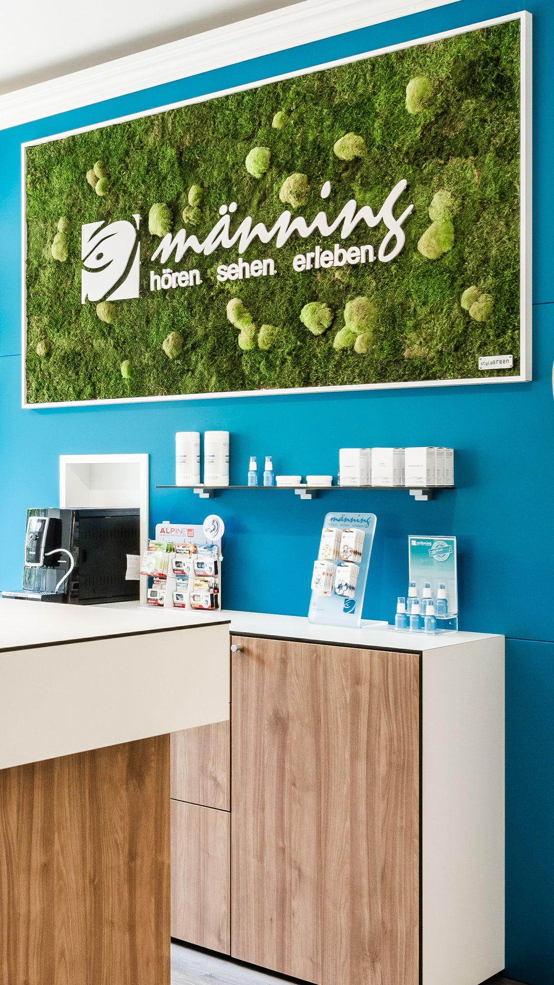 Mooswand im Männing Fachgeschäft Düsseldorf mit weißem Männing-Logo und dem Schriftzug „hören sehen erleben“.