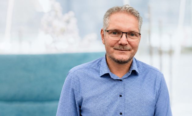 David Bruyers sitzt lächelnd in einem hellen Raum und trägt eine Brille sowie ein blaues Hemd.