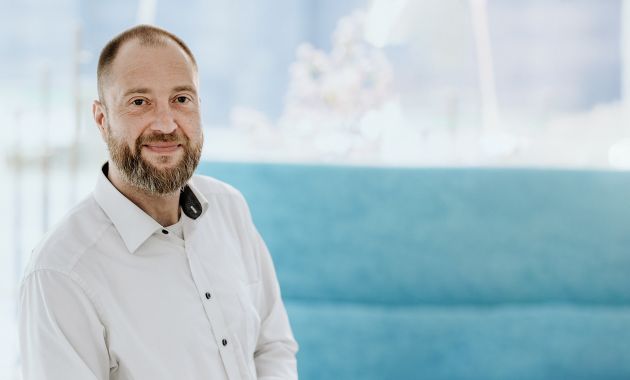 Lars Rudloff steht lächelnd in einem hellen Raum vor einem türkisfarbenen Sofa.