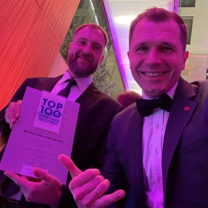 Lars Rudloff und Michael Männing sitzen lächelnd bei der TOP 100 Akustiker-Veranstaltung; Lars Rudloff hält die Auszeichnung 2023/2024 in den Händen.