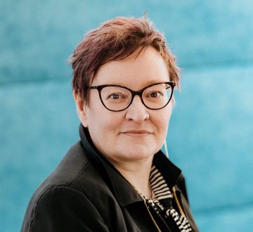 Daniela Göken sitzt lächelnd in einem hellen Raum vor einem türkisfarbenen Hintergrund.